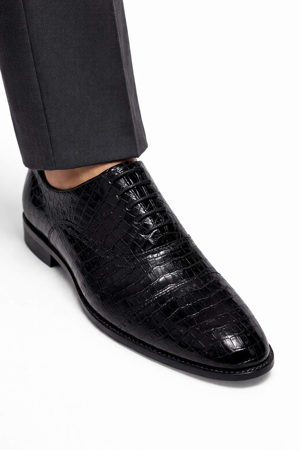 Black dress shoe with crocodile texture worn with a black suit on a white background